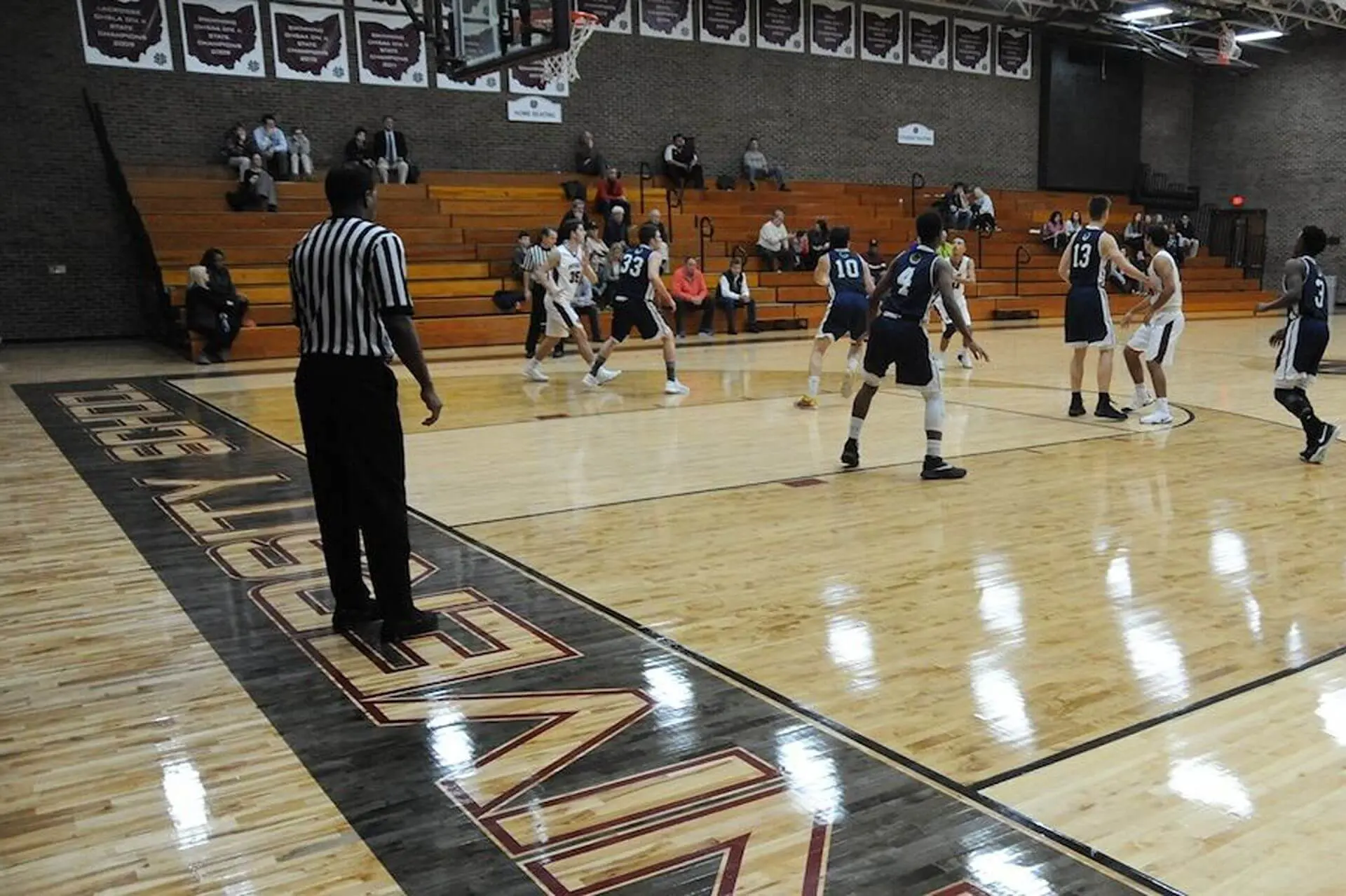 A referee in a striped uniform adjudicates over a Basketball game in the University School gymnasium.