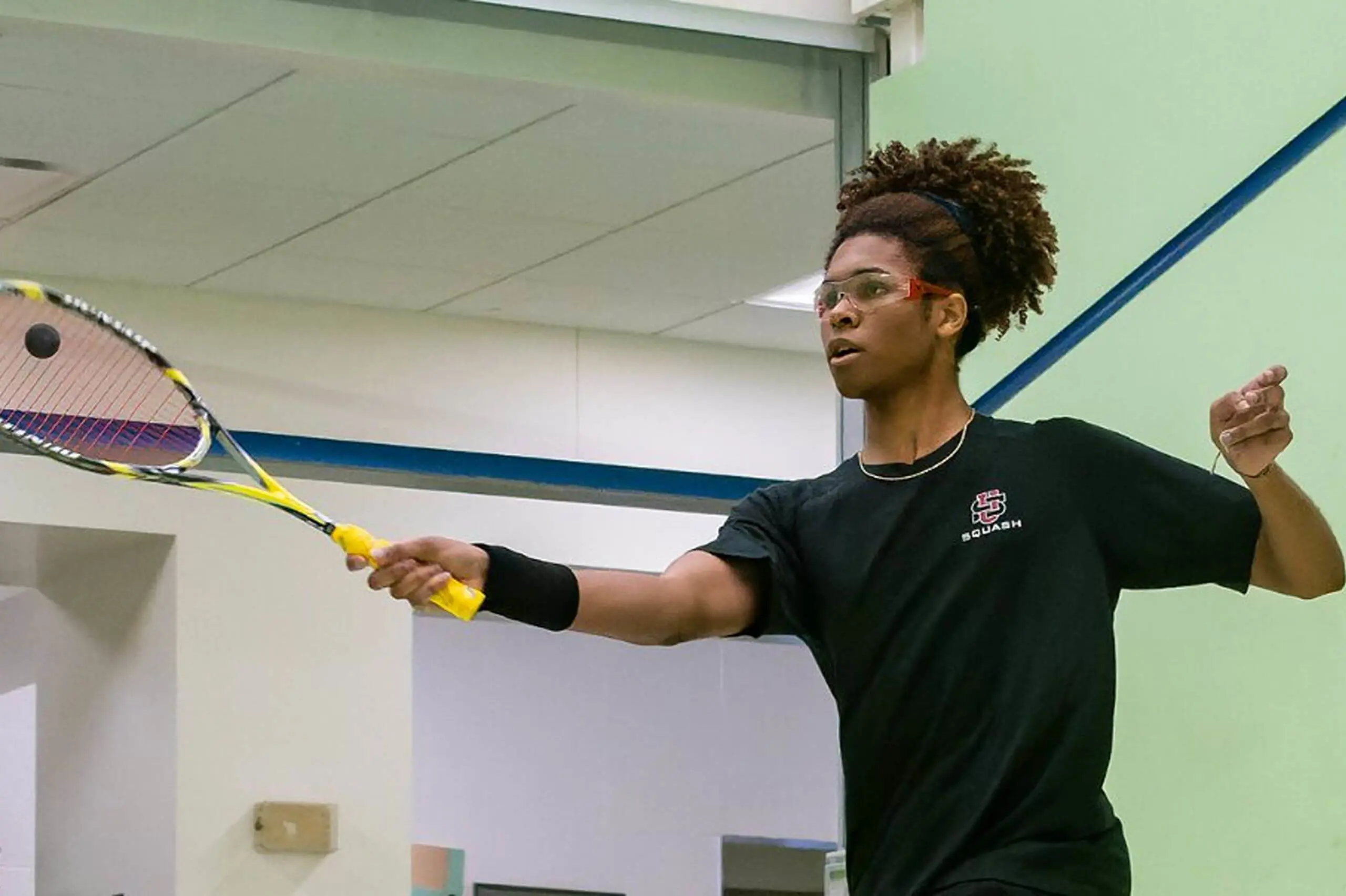 University School student wearing goggles hits a ball with a racket during a game of squash.