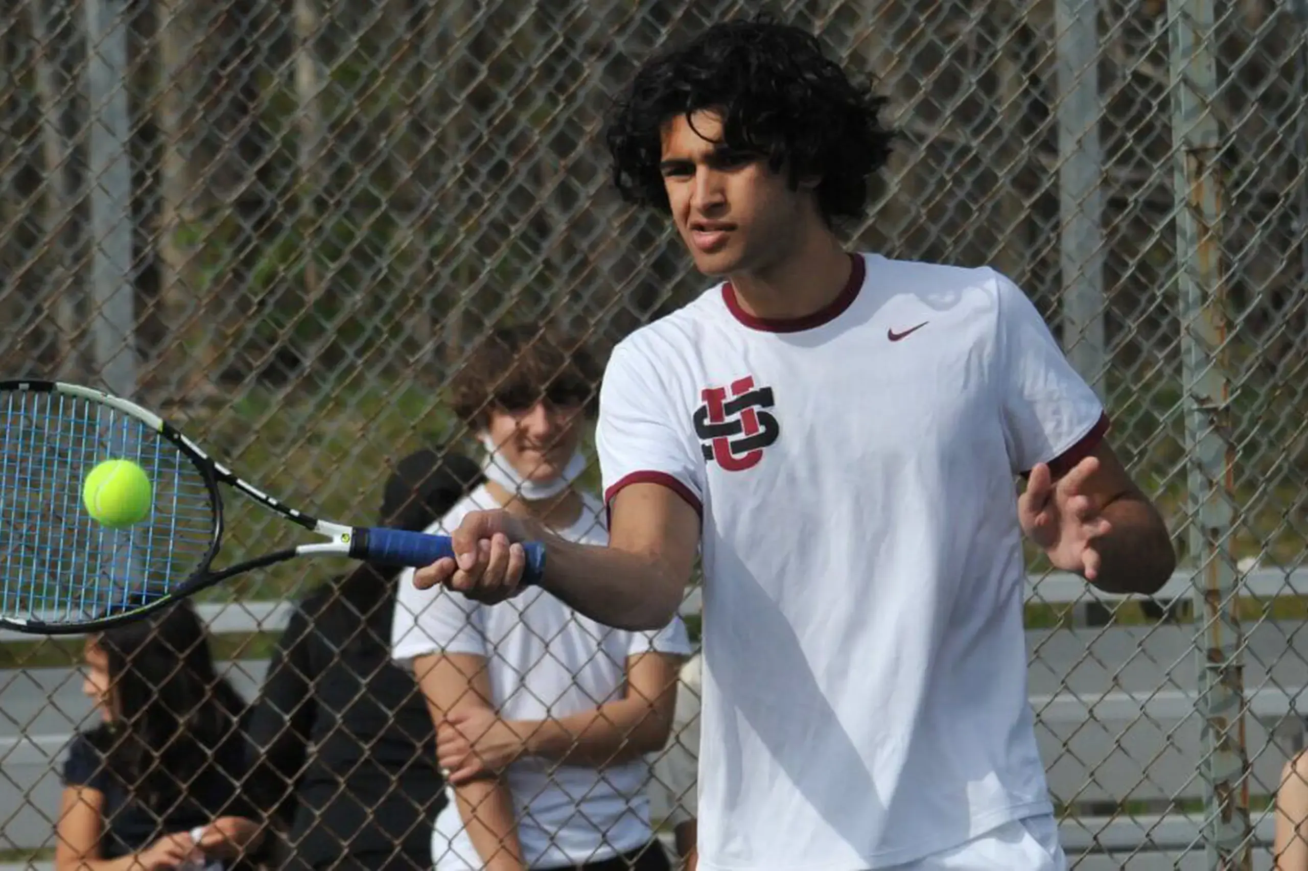 University School students hits a ball with a racket during a tennis match while other students watch.