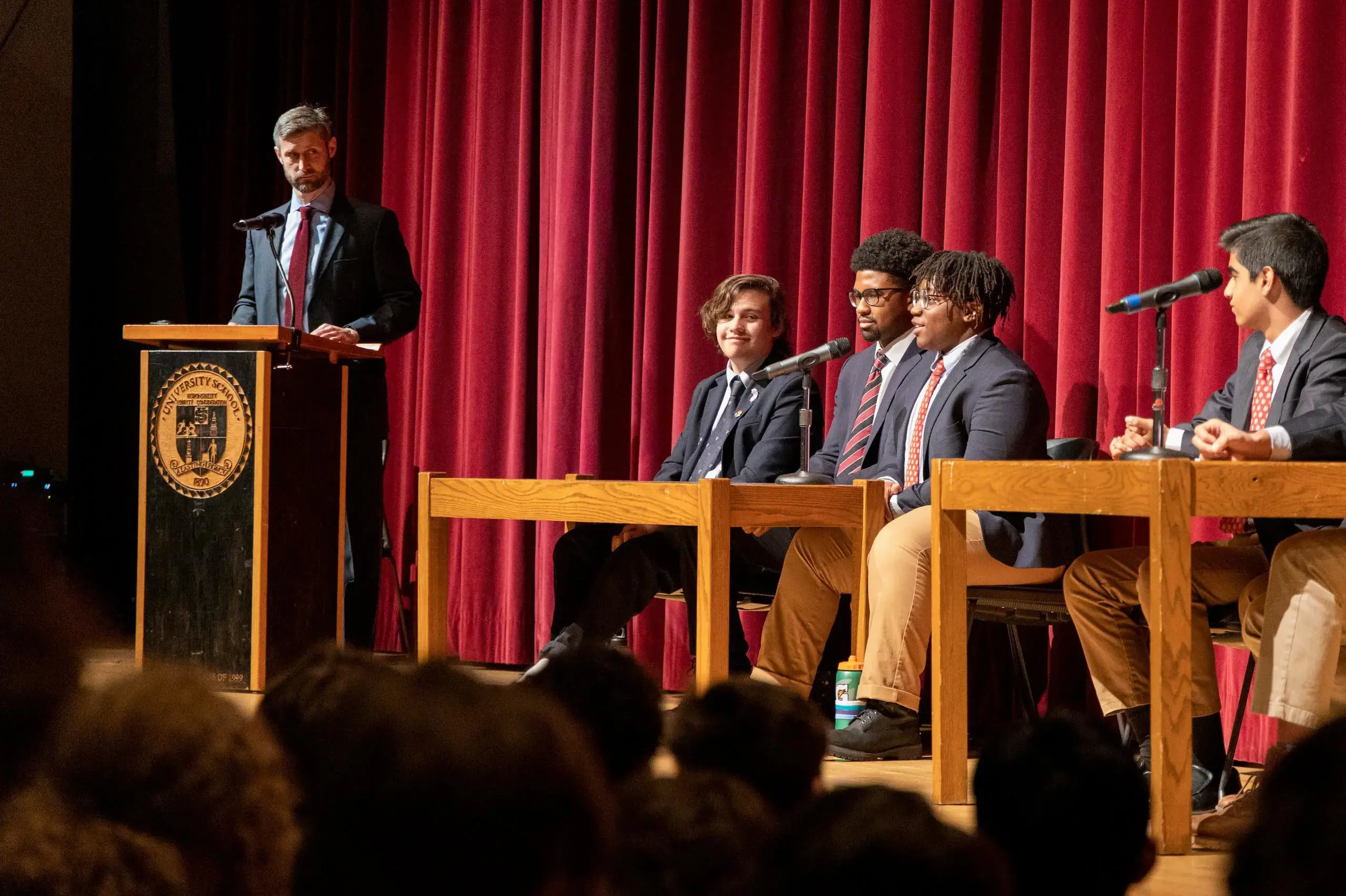 University School students holding a discussion on stage, seated at tables while a teacher looks on from a lectern with a microphone.