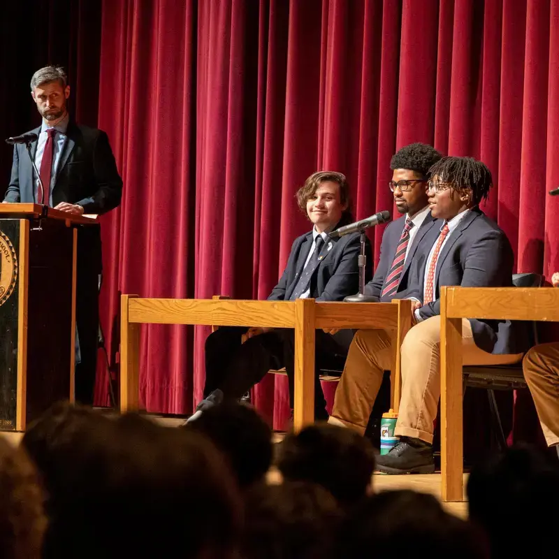 University School students holding a discussion on stage, seated at tables while a teacher looks on from a lectern with a microphone.